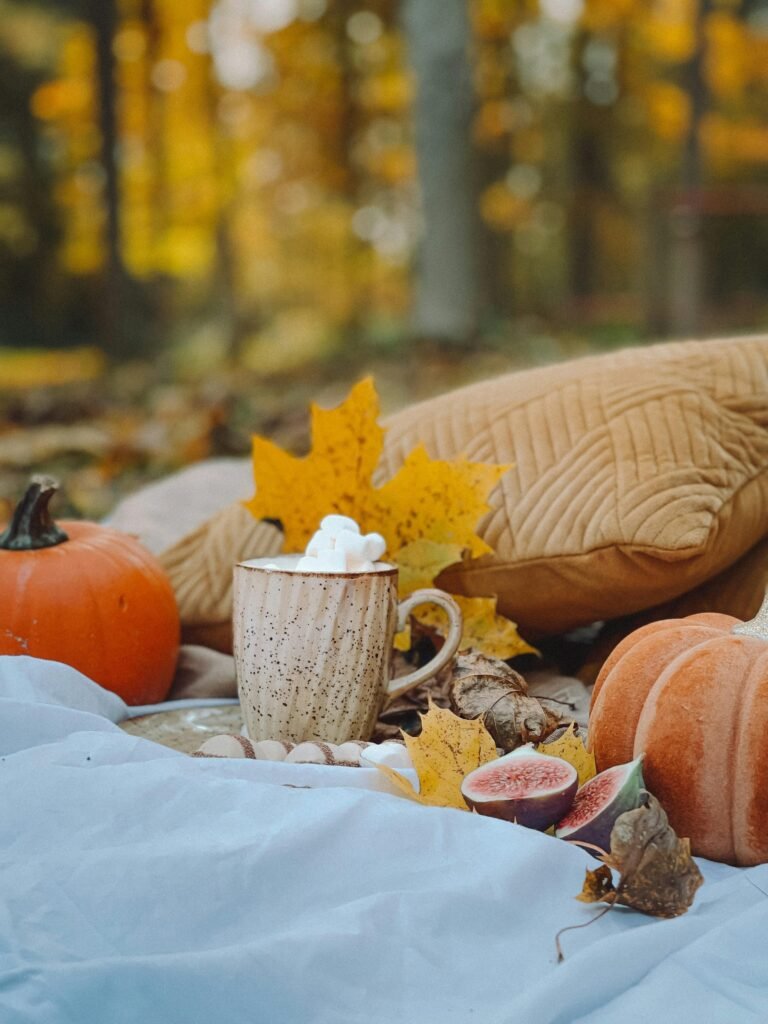 A picturesque fall picnic set up on a blanket amidst vibrant autumn leaves. The scene features a wicker basket filled with seasonal snacks, warm drinks in cozy mugs, and colorful plates. Surrounding the picnic are trees adorned with red, orange, and yellow foliage, creating a beautiful backdrop as two people sit together, laughing and enjoying the crisp fall air.