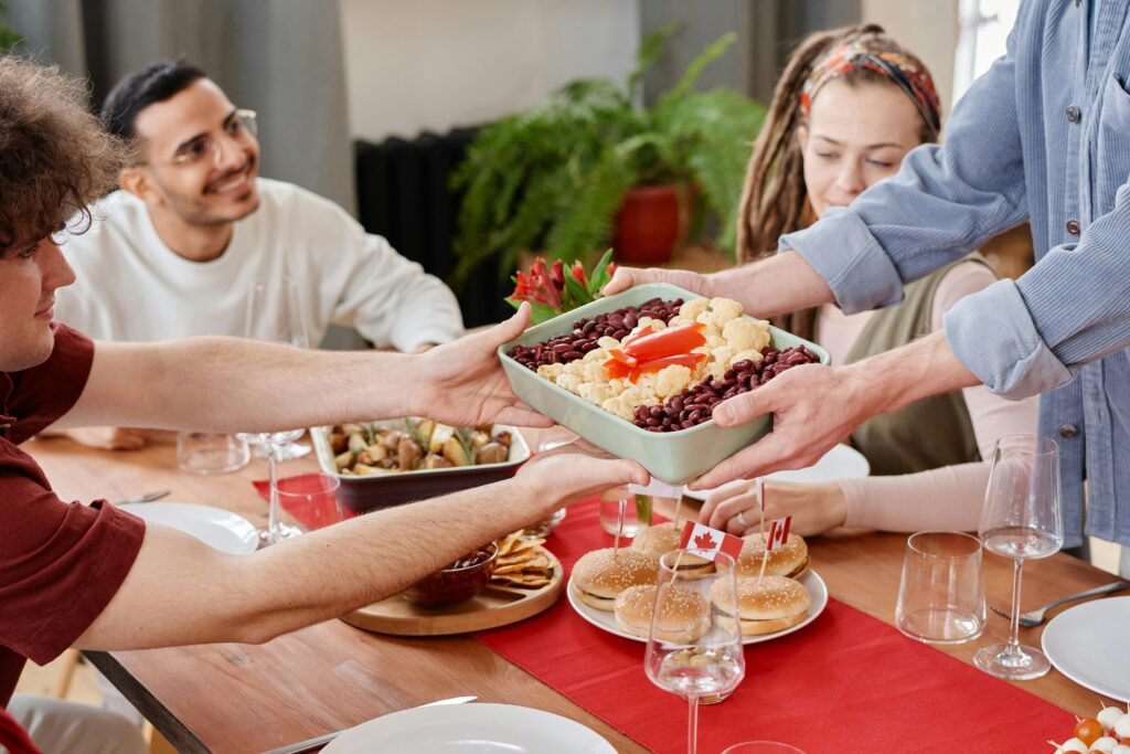 A group of people gathered around a buffet-style table filled with a variety of fall-themed dishes, including casseroles, pumpkin bread, and apple pie. The table is decorated with pumpkins, candles, and autumn leaves, creating a cozy, festive atmosphere. Guests are smiling and serving themselves, enjoying the warm and inviting potluck setting.