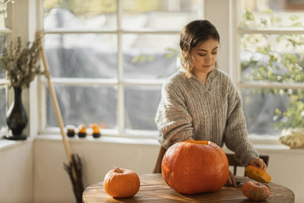 A girl focused on carving a pumpkin, using a small knife to carefully cut out a spooky face. She is sitting at a table adorned with autumn decorations, including colorful leaves and other pumpkins. The bright orange pumpkin sits in front of her, and a bowl nearby holds the pumpkin seeds and pulp. Her expression is one of concentration and joy as she engages in this festive fall activity.