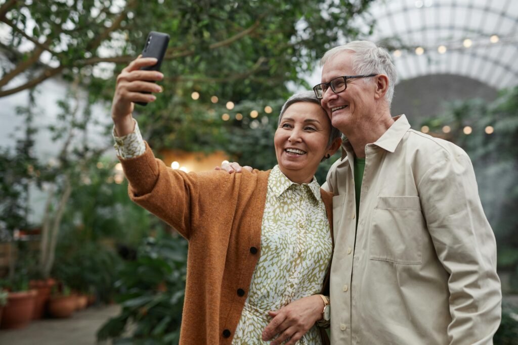 Two people smiling and posing for a picture together outdoors. They stand close, with arms around each other, against a backdrop of greenery and colorful flowers. The sunlight shines down, creating a cheerful atmosphere as they capture the moment with a camera or smartphone. Their expressions radiate happiness and friendship, highlighting the joy of sharing a special occasion.