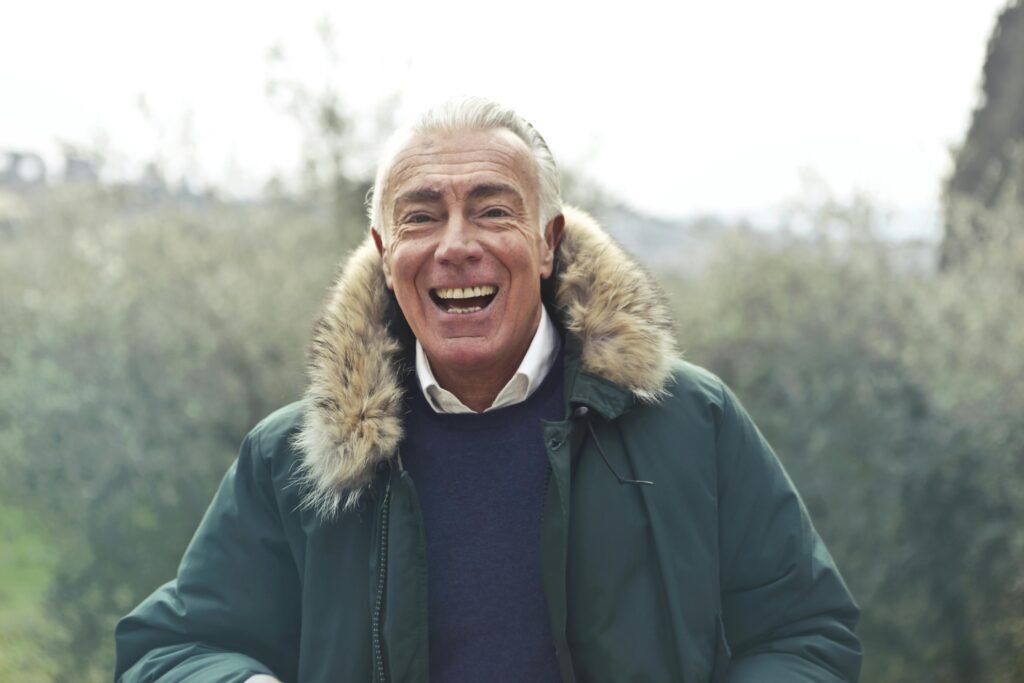 An elderly man wearing a thick, warm coat stands against a plain background, looking directly at the camera. His expression is calm and thoughtful, and the coat provides a cozy appearance, capturing the essence of staying warm in the fall chill.