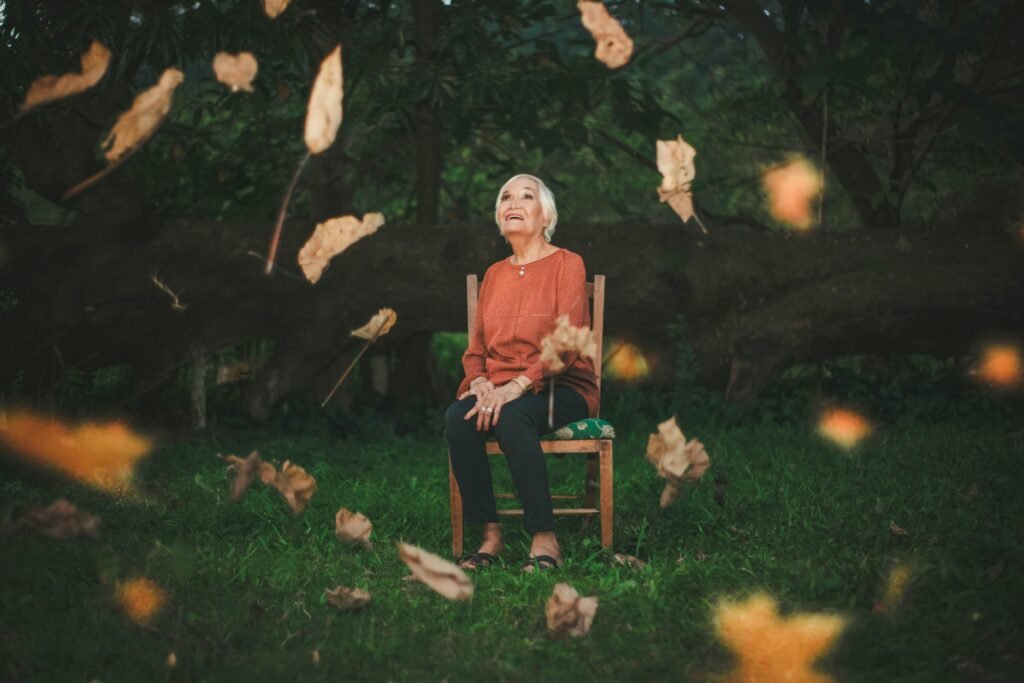 A woman sitting in a chair outdoors, looking up with a joyful expression as colorful autumn leaves fall around her. She is wearing a cozy shirt, and the vibrant hues of red, orange, and yellow leaves create a beautiful backdrop, capturing the essence of the fall season.