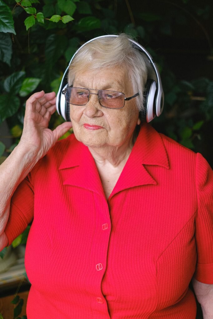 An elderly woman stands comfortably, wearing headphones and listening to music, with a serene expression on her face. She appears relaxed and fully absorbed in the moment, surrounded by a cozy atmosphere. Soft light highlights her features, emphasizing her contentment as she enjoys her favorite tunes.