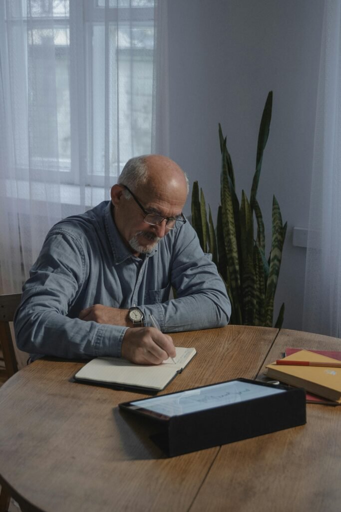 A senior man sits comfortably in a cozy corner of his home, writing in a journal. He has a thoughtful expression as he reflects on his thoughts and experiences. Soft light filters through a nearby window, creating a warm and inviting atmosphere. A cup of tea rests on a table beside him, adding to the peaceful ambiance of the scene.