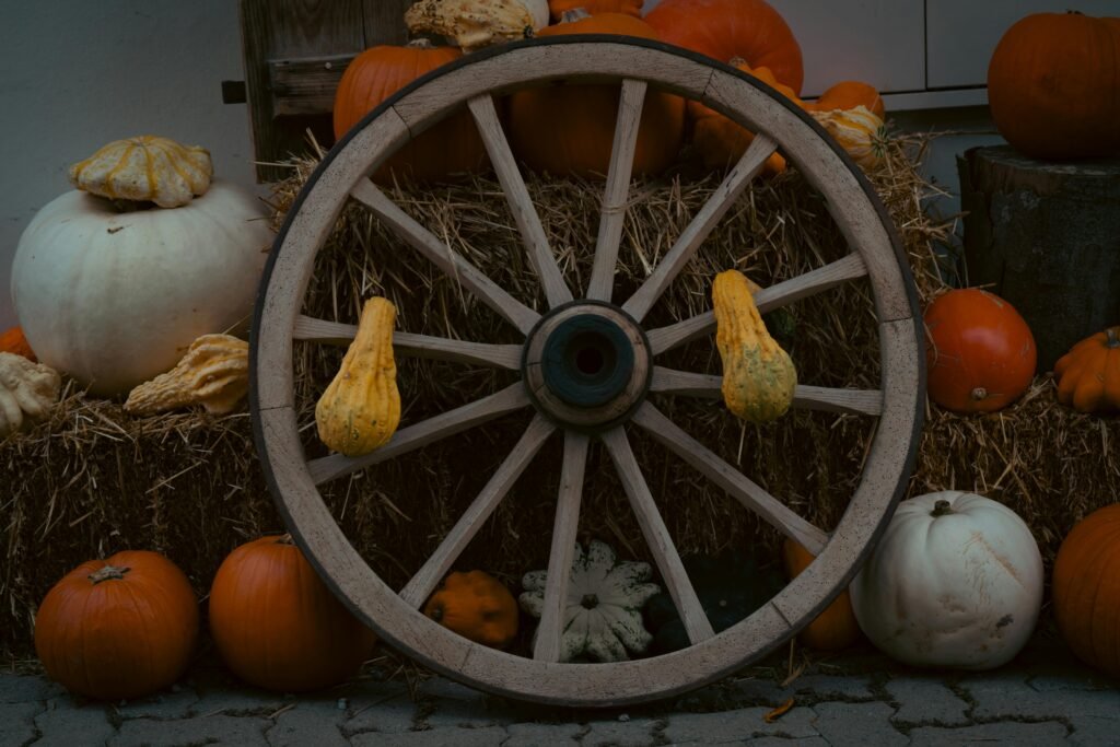 A rustic wooden wheel positioned in front of a backdrop of pumpkins stacked on bales of hay. The warm orange tones of the pumpkins contrast with the muted colors of the wheel, creating a cozy autumn atmosphere. The scene is adorned with scattered leaves, enhancing the festive spirit of the fall festival.