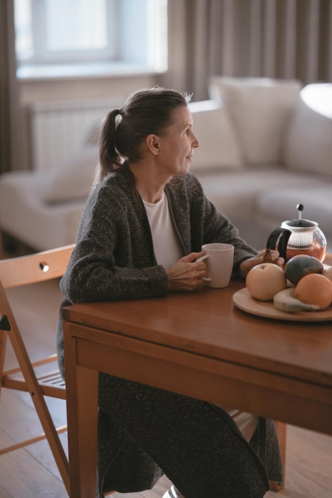 An elderly woman sits comfortably in her senior apartment, enjoying a quiet moment with a cup of coffee. She is seated in a cozy chair, surrounded by soft lighting and warm decor that creates a welcoming, peaceful atmosphere. The scene reflects a sense of relaxation and comfort, showcasing the tranquility of independent living in a senior apartment.