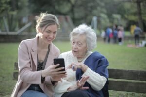 An elderly woman and her caregiver sit together on a bench in a park, enjoying the outdoors. The caregiver is smiling and gently engaging with the woman, who looks content and relaxed. The scene captures a peaceful moment of companionship, with trees and greenery in the background, highlighting the caregiver's supportive role in enhancing the woman's well-being.