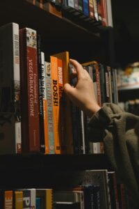 A person browsing through a selection of books on a shelf, carefully hand-picking a book to read. The books are arranged neatly, with colorful covers and titles visible, creating an inviting atmosphere for reading