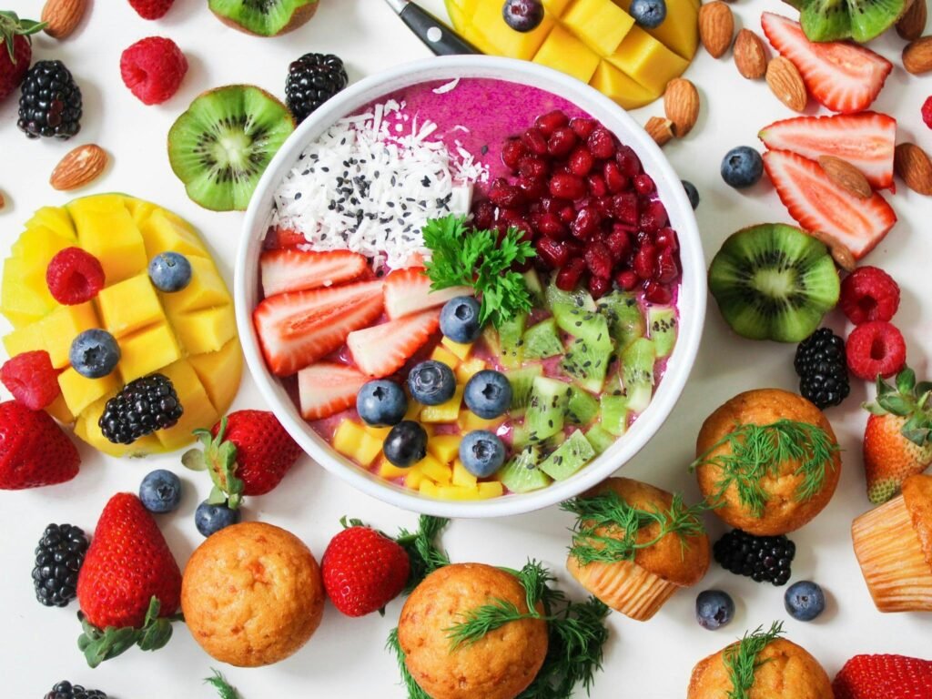 Colorful fruits in a bowl surrounded by other fruits and muffins in a white background