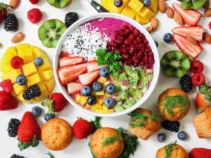Colorful fruits in a bowl surrounded by other fruits and muffins in a white background